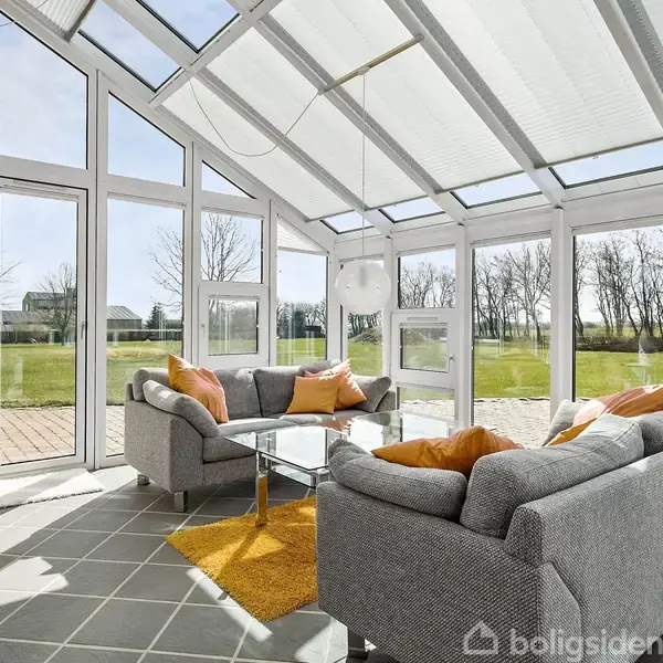 To sofaer med farverige puder står ved et glasbord i en rummelig udestue. Store vinduer og glasdøre åbner op til en terrasse og stor græsplæne.