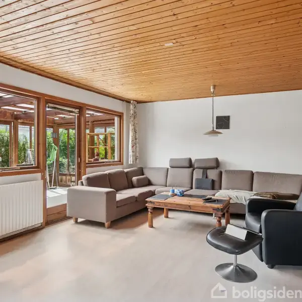 En grå hjørnesofa står i en stue med hvidmalede vægge og træloft. Ved siden af sofaen er der en sort lænestol og sofabord. Store vinduer giver udsigt til terrassen.