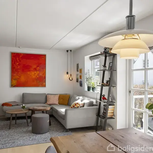 En stue med en grå sofa og farverige puder, hvor et moderne maleri hænger på væggen. Der er et spisebord med lampe og en reol ved en glasdør.