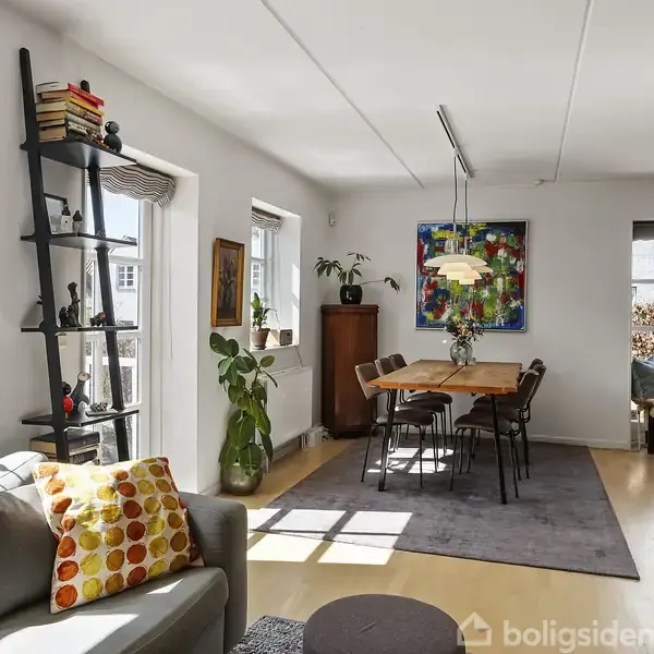 Stue med gråt sofaområde, farverig pude og reol til venstre; spisebord med stole og lampe hænger over det i midten; grønne planter og farverig kunst på væggene.