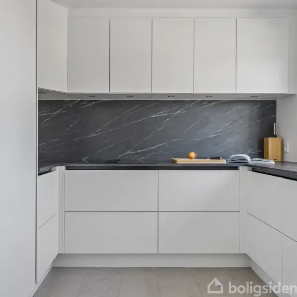 Hvidt, moderne køkken i minimalistisk design med glatte skabsfronter. Et mørkt marmorlignende bagstykke skaber kontrast. En appelsin og bog ligger på bordpladen. Vindue til højre for lysindfald.