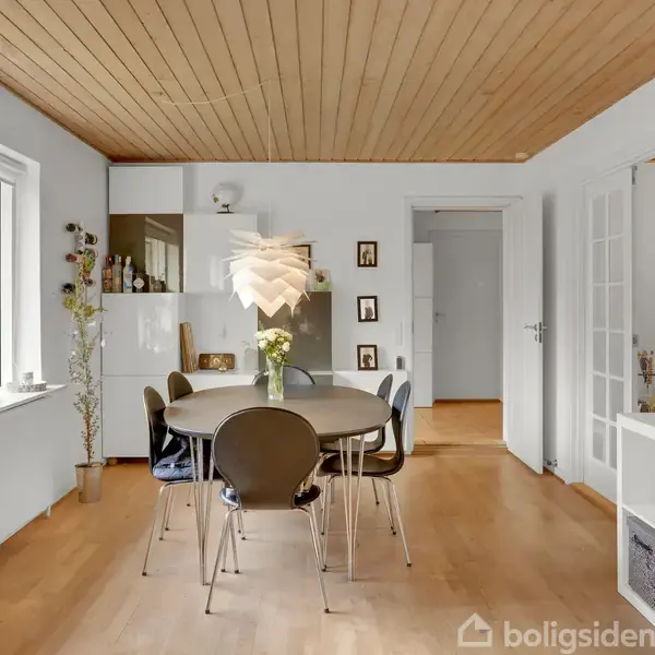 Et spisebord med seks stole står i et lyst rum med hvidmalede vægge og trægulv. En pendellampe hænger over bordet, og flere malerier hænger på væggene.
