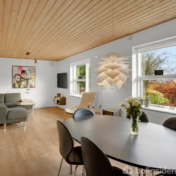 Moderne stue med lys trægulv og træloft. Rundt spisebord med stole i forgrunden, sofa og lænestol i baggrunden. Store vinduer giver udsigt til en have med buske og træer.