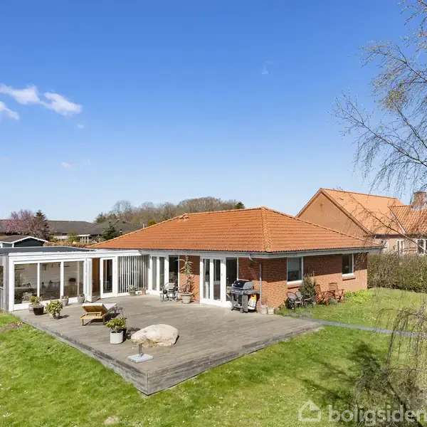 En moderne villa med rød tagstenstag står på en velplejet græsplæne omringet af terrassen. Solrige omgivelser og nabohuse kan ses i det åbne område under en klar, blå himmel.