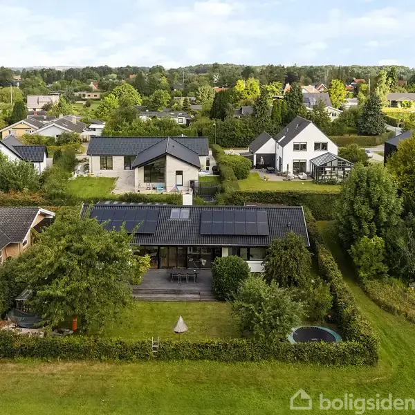 Hus med solpaneler på taget, omgivet af en have med græsplæne og træer, beliggende i et villakvarter med flere huse og grønne områder i baggrunden.