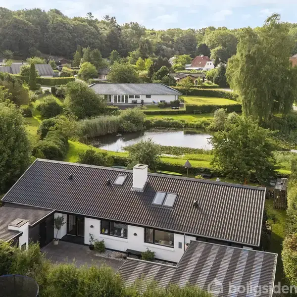En hvid villa med sort tag står i en frodig have med hæk. Omkringliggende træer og en lille sø ses, omgivet af andre huse i et grønt villakvarter.