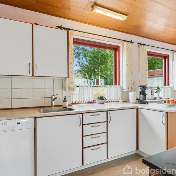 Køkken med hvide skabe og en opvaskemaskine. Køkkenvasken er placeret under et vindue med udsigt til have. Et spisebord ses i baggrunden.