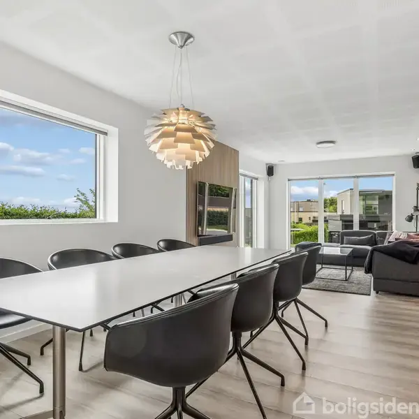 Et moderne spisestue med sort-hvide møbler. Et langt bord med sorte stole står centralt. En unik loftslampe tilføjer stil. Store vinduer giver udsigt til en grøn have.