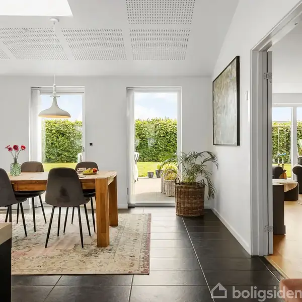 Et spisebord med stole står i et lyst rum med store vinduer, der åbner mod en have. Der hænger lamper over bordet, og til højre ses en plante.