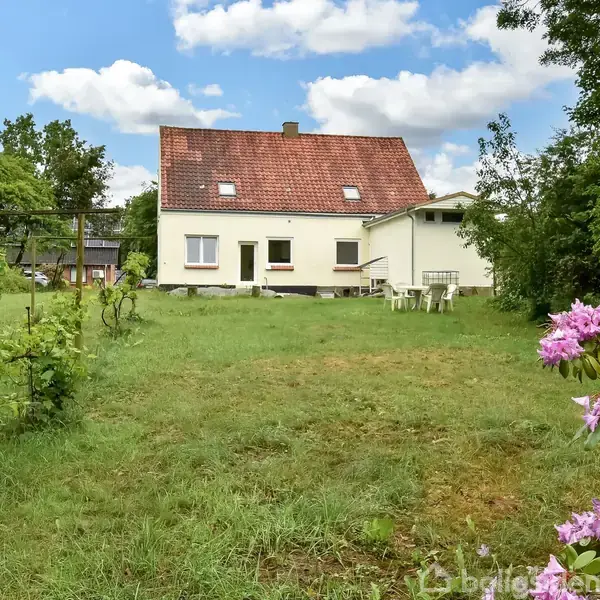 Hus med rødt tag står stille i en frodig have fyldt med græs og farverige blomster. Et bord med stole står til højre under en blå himmel med skyer.
