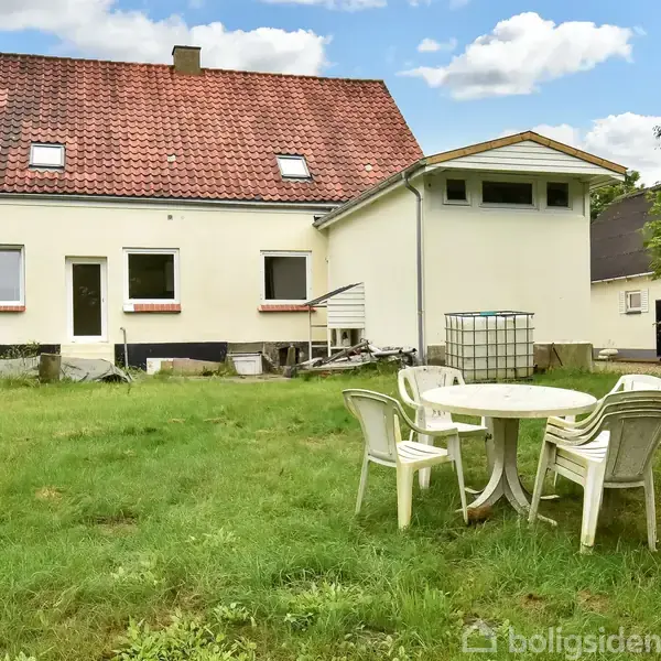 Huset står med rødt tegltag og hvid facade, omgivet af en grøn græsplæne. Fire hvide plastikstole og et rundt bord står i haven.