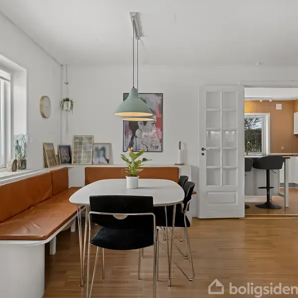 Et spisebord med stole står i et lyst køkkenalrum. Ved siden af er der en indbygget bænk langs væggen. En plante i en krukke pryder bordet, og der er adgang til køkken gennem glasdøre.