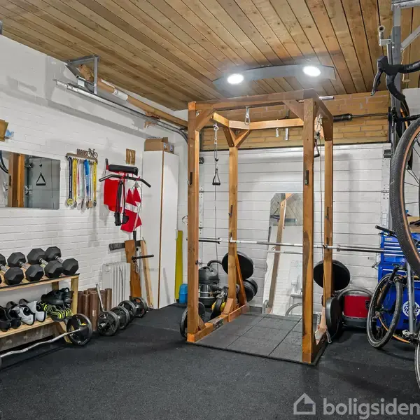 Cykler hænger på væggen, imens håndvægte er organiseret på en reol. En træningsramme står midt i rummet omgivet af vægudstyr og en spejlvæg. Loft i træ.