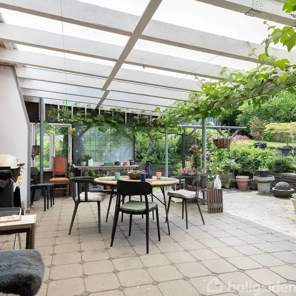 En overdækket terrasse med et bord og stole placeret på fliser. Planter og potter pryder området, omgivet af en frodig have.