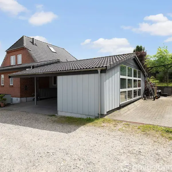 Et rødt murstenshus med gråt tag står stille ved siden af en moderne garage med store vinduer. I baggrunden er der grønne træer og buske under en blå himmel.