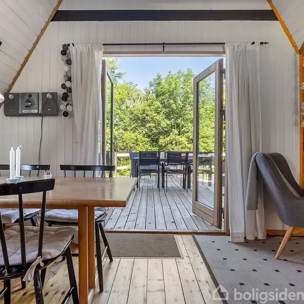 Spisebord med sorte stole står ved siden af åbne glasdøre, der fører ud til en træterrasse med borde og stole. Omgivet af grønne træer og lysindfald.