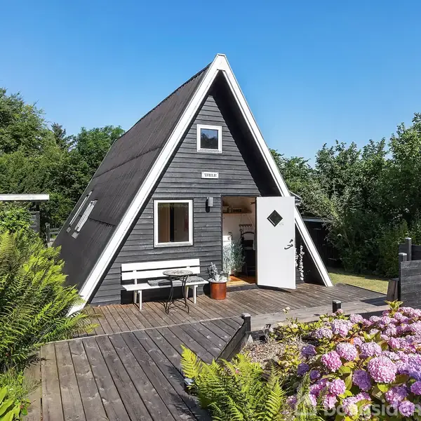 A-formet sommerhus i sort træ med åbent dør. Navneskilt "Hilltop" over døren. Omgivet af grønne buske, træterrasse, hvid bænk og lilla hortensiaer i forgrunden.