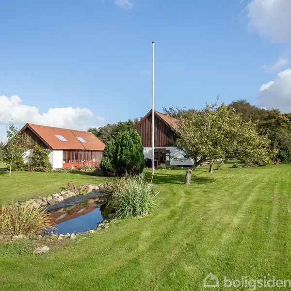 To bygninger med røde tage står i en grøn have med en lille dam i forgrunden og enkelte træer omkring. Skyfri blå himmel og spredte skyer i baggrunden.