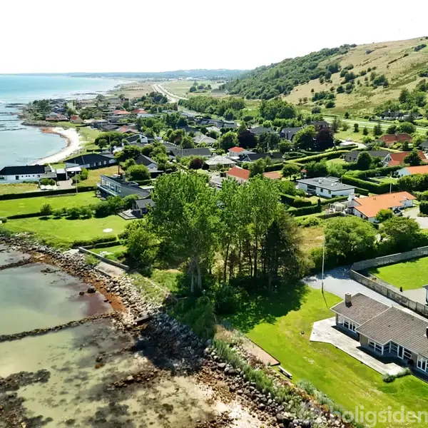 Kysthus nær havet, omkranset af grønne træer og græsplæner i et bakket, landligt område med flere huse i baggrunden og havet langs kysten.