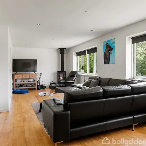 Sort læderhjørnesofa står i stue med lyse trægulve, vendt mod vægmonteret fladskærms-TV. Til venstre stor væg-ur og til højre store vinduer med udsigt til træer.