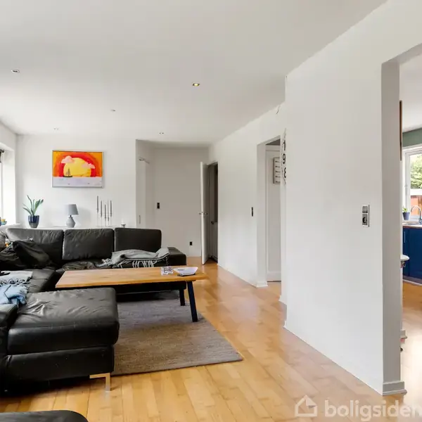 Stue med sort hjørnesofa og træbord, omgivet af lyse vægge og gulvtæppe, forbinder til køkken med blå skabe gennem en åbning, store vinduer slipper lys ind.