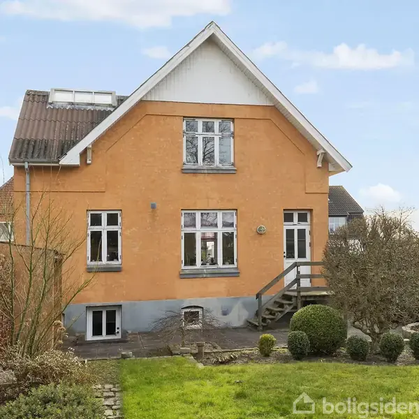 Et gulmalet hus med rødt tag og flere vinduer sidder stille i en velplejet have omgivet af buske og træer. Trappe fører til indgang. Ingen tekst fundet.