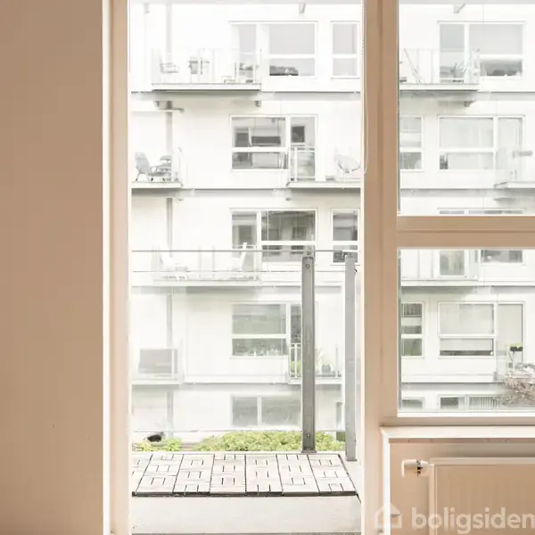 Et vindue giver udsigt til en hvid lejlighedsbygning med flere altaner. Bygningen har store vinduer og står omgivet af grønne planter i forgrunden.