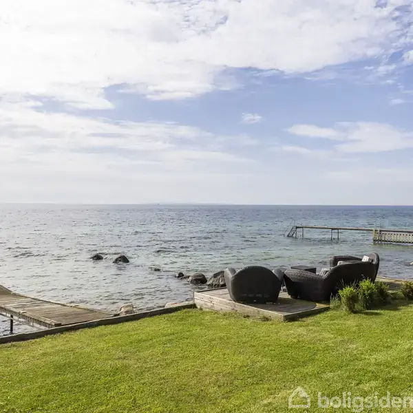 En træbro strækker sig ud i havet fra en græsplæne med sorte, runde havemøbler. I baggrunden ses en anden bro og bølger under en delvist skyet himmel.