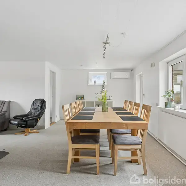 Et spisebord med otte stole står i et lyst rum med store vinduer. Til venstre er der en lænestol og en kamin. Rummet virker rummeligt og velbelyst.