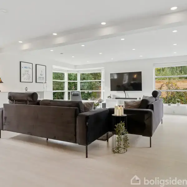 To sorte sofaer står i en lys stue med store vinduer. Et fjernsyn hænger på væggen, mens lamper og planter tilføjer hygge til rummet.