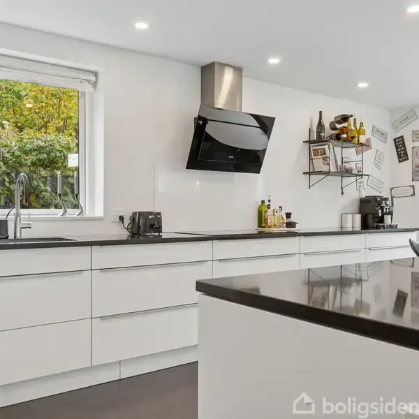 Køkken med hvide skabe og sort bordplade. Emhætte og hylder med dekorationer. Vindue viser grønt løv udenfor. Lyse vægge og gulv. Sollys trænger ind i rummet.