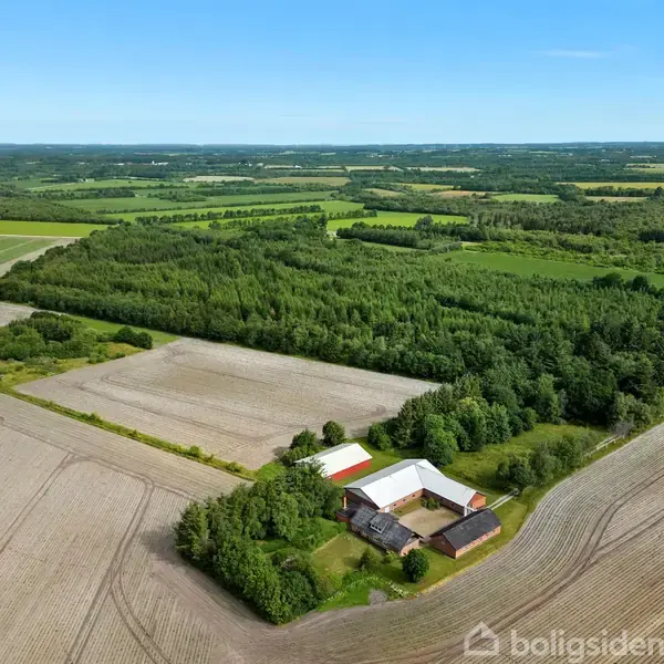 En stor gård med flere bygninger ligger midt i et landskab af marker og skovområder under en klar blå himmel. Tekst: "boligsiden".