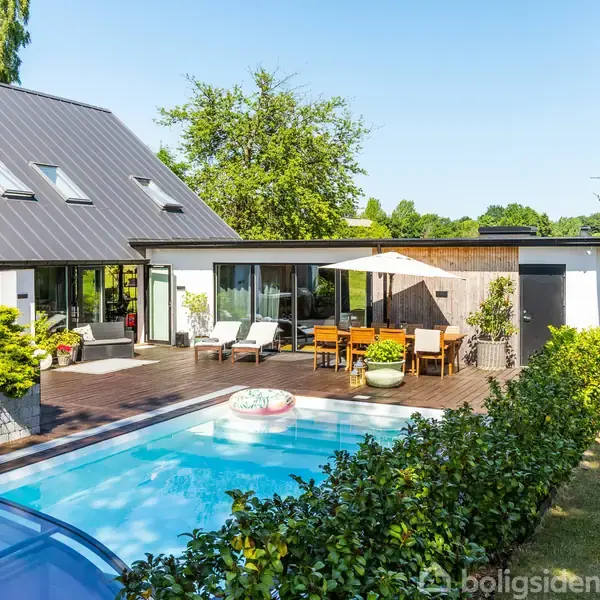 Hus med sort tag ligger ved siden af en swimmingpool. Terrasse med liggestole, spisebord og parasol omkranser poolen. Grønt haveområde i baggrunden under blå himmel.