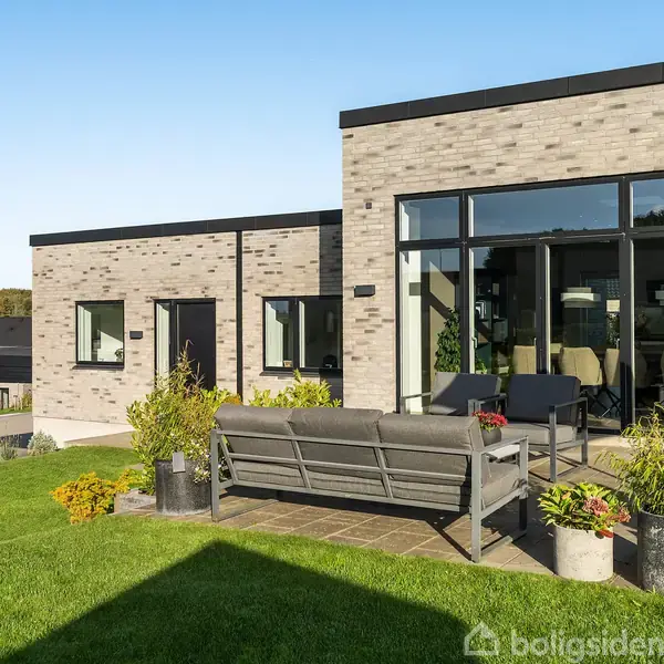 En moderne terrasse med en grå sofa og borde står på fliser foran et hus i grå mursten. Grønne planter omkranser området, og blå himmel ses i baggrunden.
