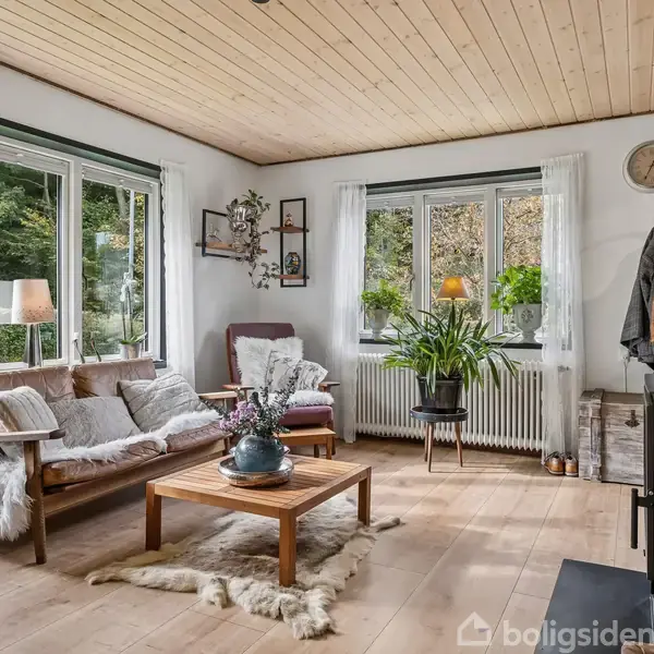 En hyggelig stue med en brændeovn i hjørnet, varm ild i ovnen, en sofa og lænestol med tæpper, træbord i midten, og store vinduer mod grønne omgivelser.