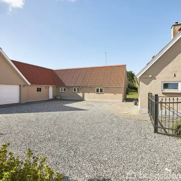 Gårdhus med rødt tegltag og beige facade står i solrigt vejr. Grusbelagt indkørsel fører til husets indgang. To hvide garageporte til venstre, grønt område i baggrunden.