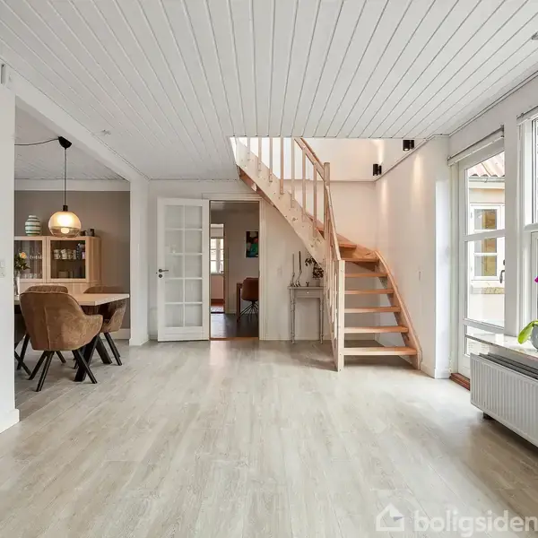 Trætrappe fører opad i lyse stue med trægulve, hvide vægge og loft. Store vinduer til højre lukker dagslys ind. I baggrunden er spisebord med lamper over og skænke.