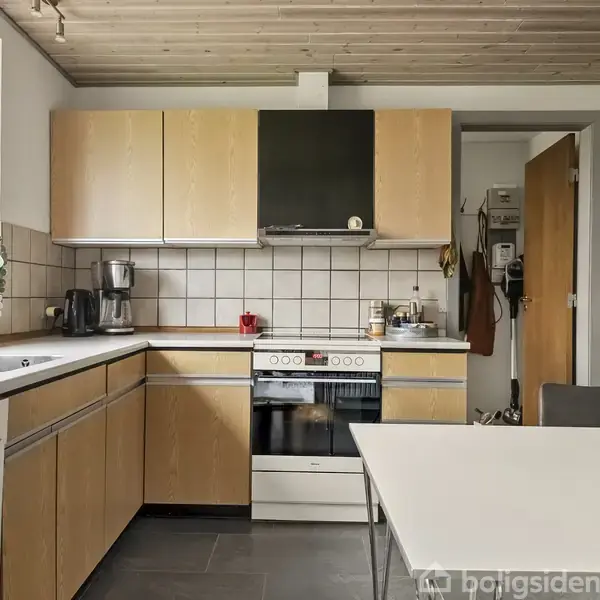 Køkken med træskabe og hvide fliser over komfuret. Bordflade ved siden af en opvaskemaskine. En bordplade med kaffemaskine og planter. Bord med stol i forgrunden.