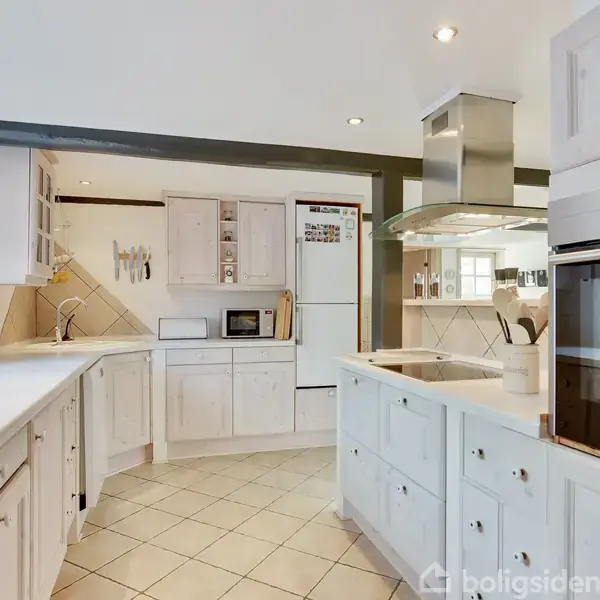 Et moderne køkken med hvide skabe og apparater, inklusive ovne og mikroovn. Det har en central ø med indbygget kogeplade og emhætte. Indretningen er lys med flisegulv.