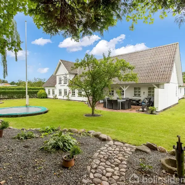 Hvidt hus med skråt tag står på en velplejet græsplæne omgivet af træer. I haven er der en flagstang, trampolin og terrasse med havemøbler.