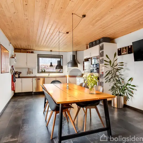 Et spisebord står i et køkken med træloft og mørkt stengulv. Bordet er omgivet af stole, og en lampe hænger over det. Der er planter og et tv på væggen.