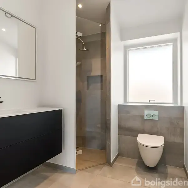Et moderne badeværelse med toilet og vask. Vasken har et sort skab, og der er et spejl over den. En bruseniche findes til venstre, og rummet har lyse vægge og gulv.