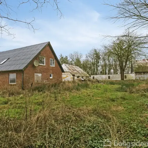 Rødt murstenshus med solcellepaneler på taget står ubenyttet. Græsset er tilgroet i en landlig have omgivet af træer. I baggrunden ses flere gamle bygninger.