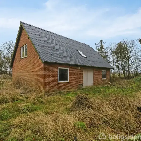 Rødt murstenshus med sort tag står stille i en overgroet have med græs og buske. Omgivet af høj trævækst under en fortrinsvis blå himmel.