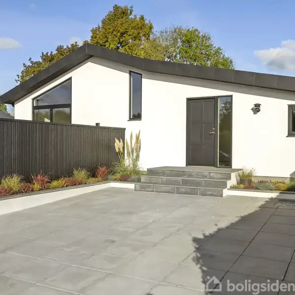 En moderne, hvid villa står stille med sort tag og vinduesrammer. Foran er en flisebelagt gårdsplads med trin til hoveddøren, omgivet af en sort træhegn og planter.