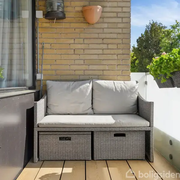Grå sofa med hynder står på en træterrasse. Den er omgivet af en murstensvæg med blomsterkrukker og planter i altankassen, hvilket skaber en hyggelig og grøn atmosfære.