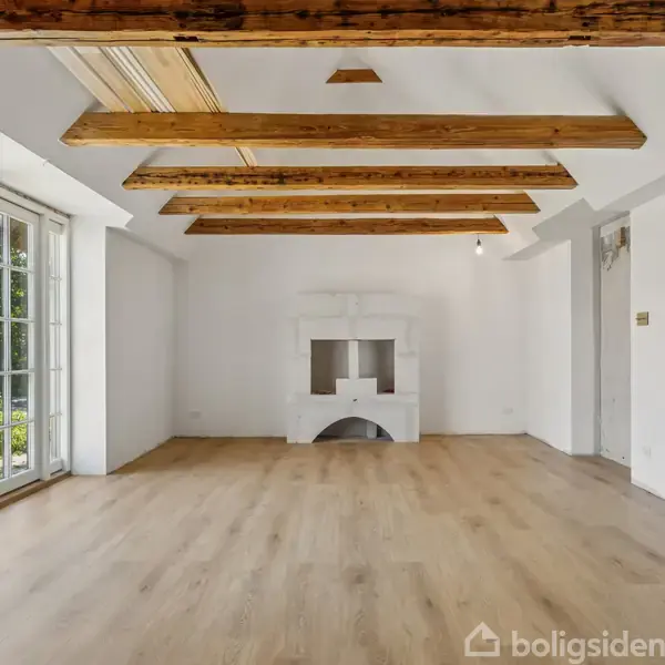 Et hvidt rum med trægulve og synlige træbjælker i loftet. Rummet har en hvid pejs i midten, og store vinduer til venstre, der viser en grøn have udenfor.