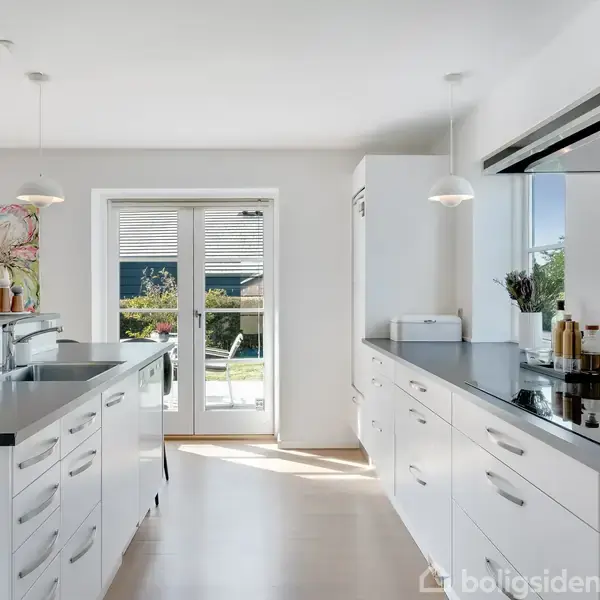 Lyst køkken med hvide skabe og sorte bordplader, centralt placeret køkkenø med vask, vinduer og døre i baggrunden giver udsigt til have. Maleri hænger over bordpladen.