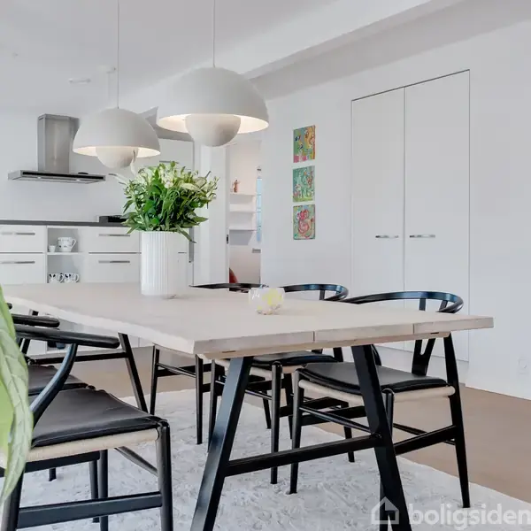 Et spisebord med sorte stole står i et lyst køkken. En hvid vase med blomster pryder bordet. Over bordet hænger store, hvide lamper. Malerier hænger på en hvid væg.