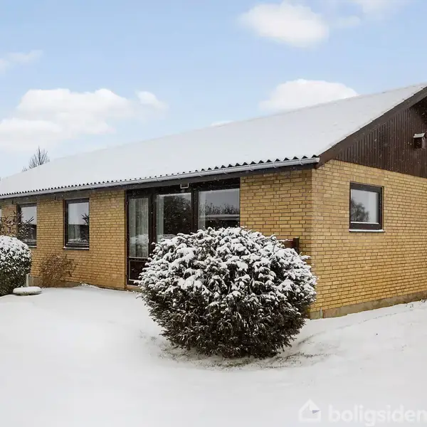 Et gult murstenshus med sort tag står dækket af sne. Foran huset er der en snedækket busk. Træer og en snedækket græsplæne udgør den omkringliggende have.